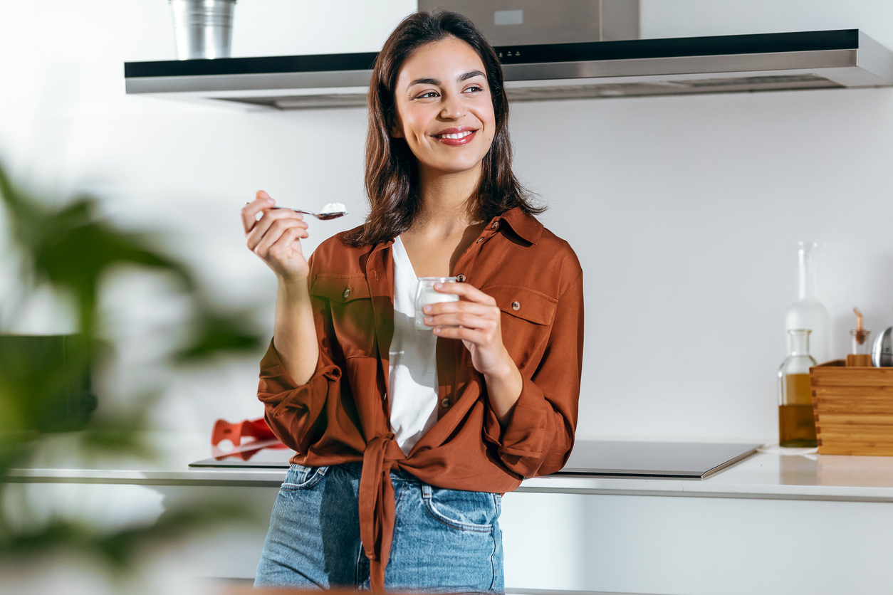 Beautiful happy woman eating a yoghurt while looking forwards in the kitchen at home